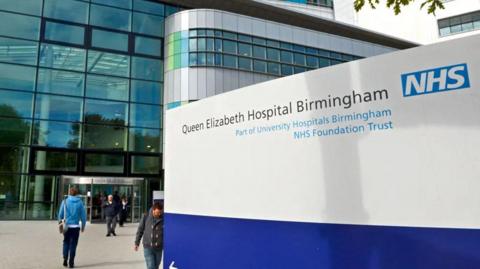 A large blue and white sign which reads Queen Elizabeth Hospital Birmingham in front of the glass entrance to a building with people walking outside