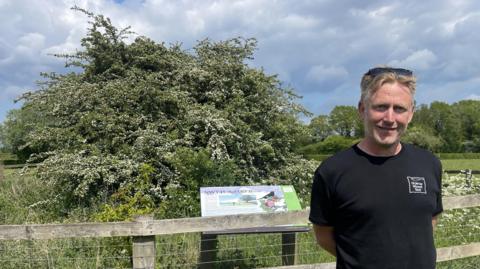 A man with a black T-shirt and blond hair smiles at the camera. He is standing in front of a tree which is behind a fence. An information panel is just behind the man but is not readable.