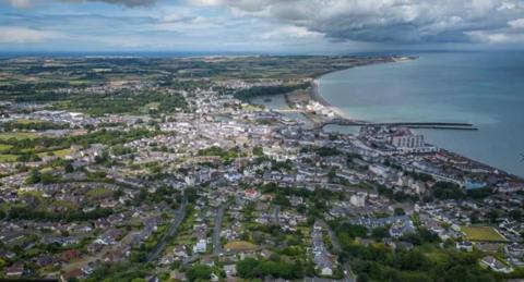 Ramsey from above with the Irish Sea to the right. Housing and a network of roads can be see beside a curving bay of blue water with a two-pronged pier stretching out into the water and clouds in the sky above.