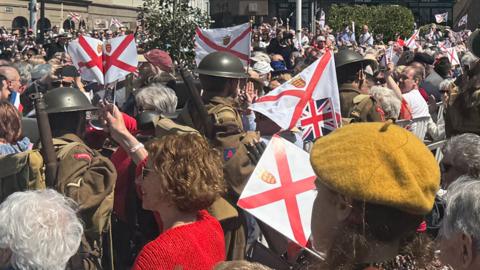 Re-enactors dressed as WW2 soldier walk through a crowd waving flags