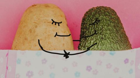 A potato and avocado with a drawn on eyes, mouths and hands under a blanket holding hands. 