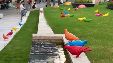 Brightly coloured sculptures of pigeons placed near water and grass in a modern, urban street in Shanghai, China