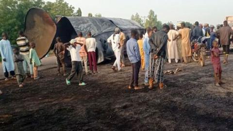 People standing around at the scene of the accident with the remains of the tanker in the background. The earth is charred.