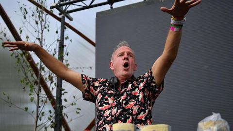DJ Fatboy Slim plays on stage at Glastonbury Festival. He is stood in front of a darkened screen and has his hands in the air with his mouth open. He has short grey hair and is wearing a black shirt with orange and white koi fish on it. He has a pair of headphones around his neck.