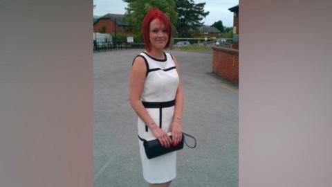 Tiffany Render has dyed-red hair and is smiling. She is standing near a building, dressed in a smart long white and black dress, holding a black clutch handbag in both hands.