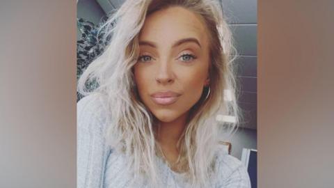 Natasha Gove, a woman with wavy, shoulder-length blonde hair, looks directly at the camera. She is wearing a light grey ribbed jumper.