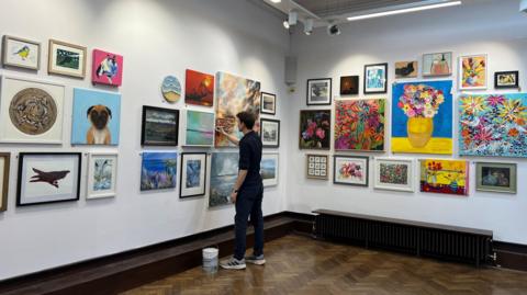 A man standing in a gallery space with an array of colourful artwork on the white walls. Among them is a picture of some brightly coloured flowers in a vase, as well as an image of a brown and white dog.