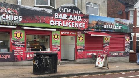 The outside of a café with a colourful fascia featuring images of Old Trafford stadium. The boards advertise food and drink and read 'United Cafe Bar and Off Licence'. 