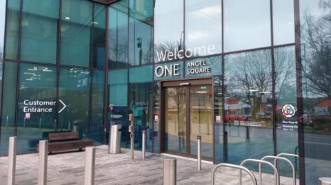 A glass fronted building with "Welcome One Angel Square" written above the doors.