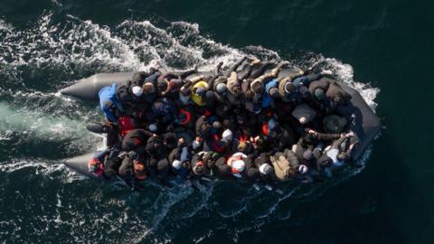 An aerial shot of a black boat at sea. It is full of people wearing coats and hats, many sat on the boat's edge.