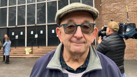 An older man wears a flat wool hat, tinted glasses and stands in front of a large brick building. 