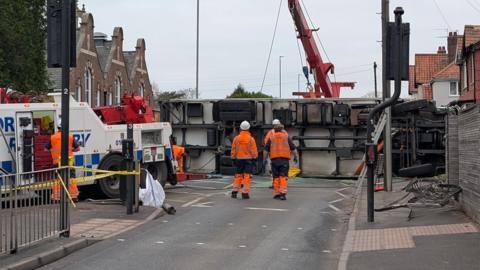 An overturned lorry on its side on a residential street. A recovery team are pictured surrounding the vehicle.