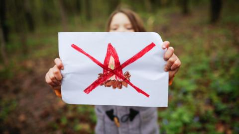 A girl holds up a white piece of paper with a hand drawn camp fire and a big red cross through it