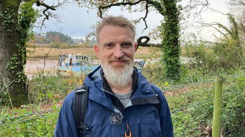 A man with a grey beard, and a blue walking jacket , stood in wooded area, with boat behind him , stranded on a mud bank