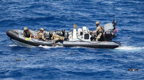 A rigid-hull inflatable boat in the sea surrounded by about seven large packages floating in the water. There are four people on the boat dressed in military camouflage uniforms and helmets. One of them is leaning over the side of the boat and pulling out one of the packages