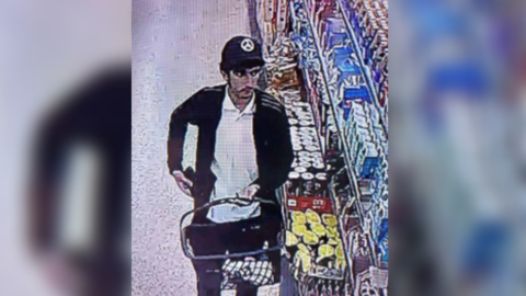 A CCTV image of a man in a black cap and jacket carrying a shopping basket