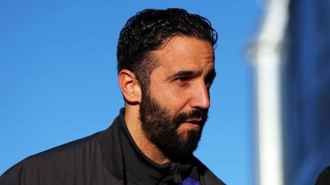 Side view of Ruben Amorim. He has short black hair and a beard, and wears a black sports jacket
