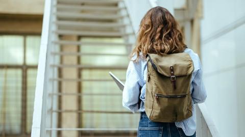 A student wearing a blue and white shirt walks away from the camera up some stairs, with a green rucksack on and carrying a laptop