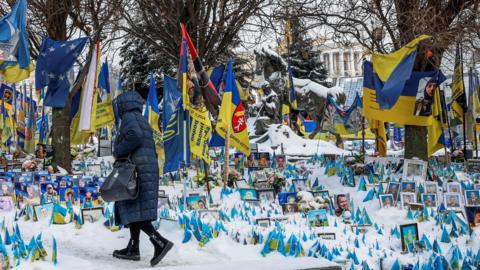 A woman dressed in an ankle-length winter coat walks at the snow-covered makeshift memorial to fallen Ukrainian personnel at Independence Square in Kyiv on a frosty winter day.