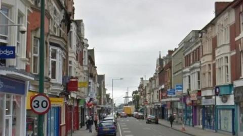 A high street with shops and parked cars dotted on either side of it with a 30mph speed limit sign seen on the left.