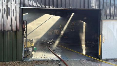 A large silver building with widespread fire damage. The upper sections of the exterior walls have been blackened by smoke. The wide entrance is open, revealing a charred interior. Fire hoses lie across the wet ground leading inside, where two firefighters are working.