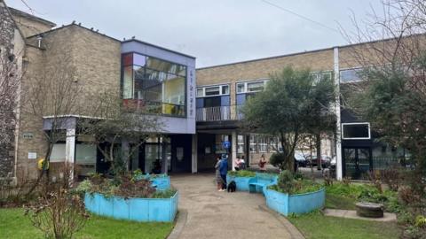 Great Yarmouth's central library, with a pathway, bordered by plants in "boat" shaped, blue raised beds, and community garden on the approach. The building features a large box window above the entrance. A few people are standing outside.