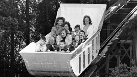 A black and white photo of children and adults in a large cart going down tracks. They are screaming in excitement.