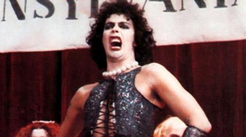 Tim Curry in a scene from the Rocky Horror Picture Show, wearing a black corset. He is singing in front of a Transylvania banner.