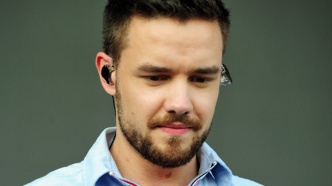 Liam Payne, with dark hair and a beard, is looking down and wearing an earpiece in front of a plain background.