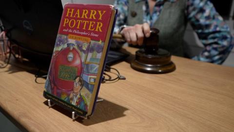 A first edition copy of Harry Potter and the Philosopher's Stone propped up on an auction table. A woman holding a gavel sits behind it. 