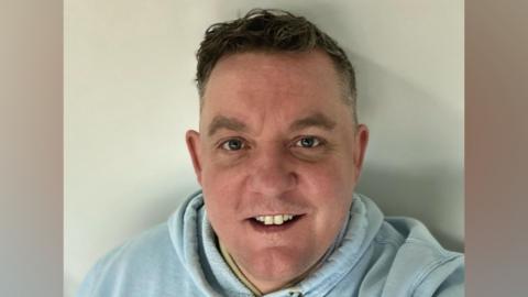 Alex Duffy has short brown hair and blue hoodie. He is smiling at camera.