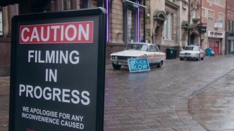 Two vintage Rover police cars, white with a red stripe across the side and blue light on top, parked up outside a row of grand-looking early 20th Century buildings, including the former Bob Carver's chip shop. A sign reads "police slow". In the foreground, another sign reads "caution, filming in progress, apologies for any inconvenience caused".