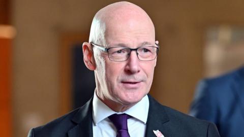 John Swinney, a bald man with glasses and wearing a dark suit with purple tie, in the Scottish Parliament