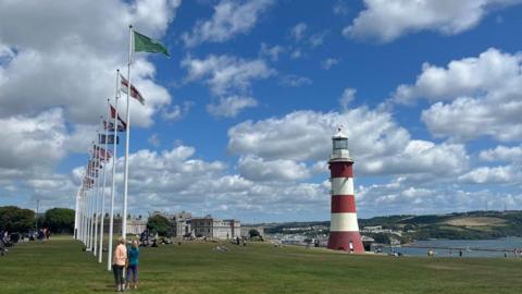 A row of flagpoles on Plymouth Hoe on the left with Smeaton's Tower, a red and white striped lighthouse on the right with the Hoe falling away to the sea.