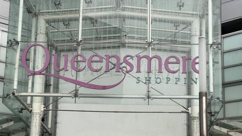 The sign of Queensmere shopping centre. 'Queensmere' is in a purple colour and 'shopping' is in a blue coral colour. 