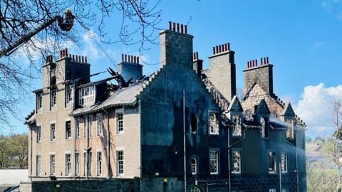 Fire-damaged hospital building in daylight, with crews using height apparatus.