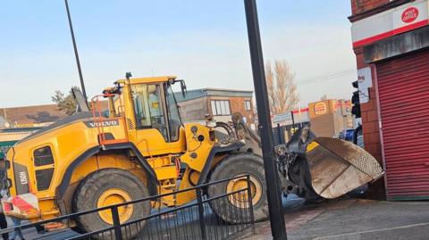 A yellow digger up against a post office, it appears to have a knocked a traffic light down