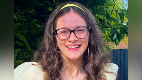 A headshot of Rosie, smiling. She has long brown, curly hair. She wears a yellow alice-band in her hair and black glasses. Her top is pale yellow and has puffy shoulders. 