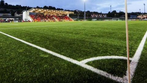 Image of the Ryan McBride Brandywell Stadium. A corner flag is visible near a large green pitch with a half-full stadium in vision. 