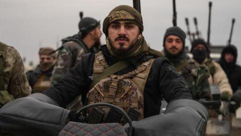 Syrian troops stand together as one soldier sits on a motorcycle, wearing tactical gear and surrounded by others carrying weapons.