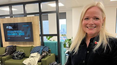 Elisabeth Lewis-Jones, chief executive of Liquid, stands near a TV screen and a green sofa with coats and bags on it. She has long blonde hair and is wearing a buttoned-up black jacket.