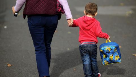 A young boy wearing a red jumper and jeans is holding a woman's hand as they walk side by side. The small child is holding a blue Minions backpack in his other hand. The woman is wearing a purple jacket and jeans. She is holding keys in her other hand.