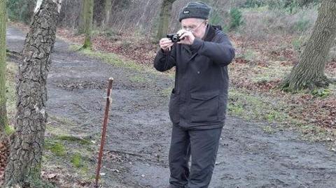 Fraser Tuddenham taking a photo of a stick in the ground with a digital camera with woodland around him. He is wearing a hat, coat and glasses.