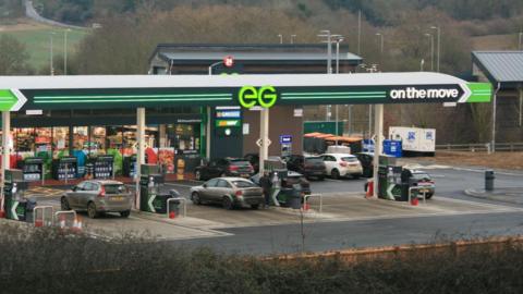 A filling station forecourt seen from a wide angle, with cars at pumps, a shop beyond and EG On the Move written across the forecourt roof