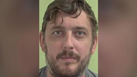 A mugshot of Liam Dunne, a man with mouse brown hair and stubble, there is a grreen background.