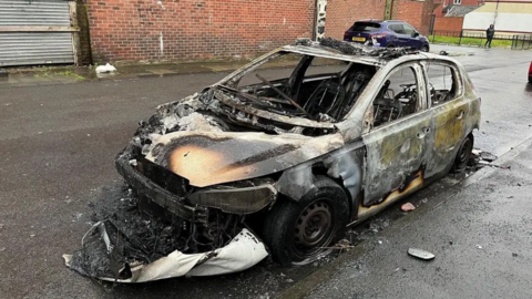 A completely burned out police car with windows fully knocked out. Ashes on the ground. 