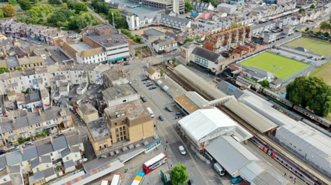 The image shows an aerial view of a busy urban area and a train station. The station has multiple platforms and tracks, with several trains visible. Nearby, there’s a bus terminal with several buses.