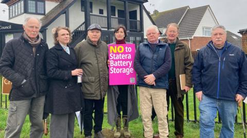 Seven people stand in a row outside a council office, in protest against the proposed battery site. A lady in the middle holds a bright pink sign saying "NO to Stater Battery Storage". There are two women and five men, all wearing winter coats.