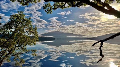 Beinn Resipol reflected in Loch Sunart. The sky is bright with patches of cloud that are reflected into the loch. Trees frame the mountain and the sun sits in the top right corner.