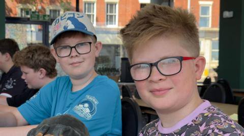 Brothers Oliver and Edward aged 13 and 11. The boys both wear glasses and are sitting next to each other at a board game cafe. 
